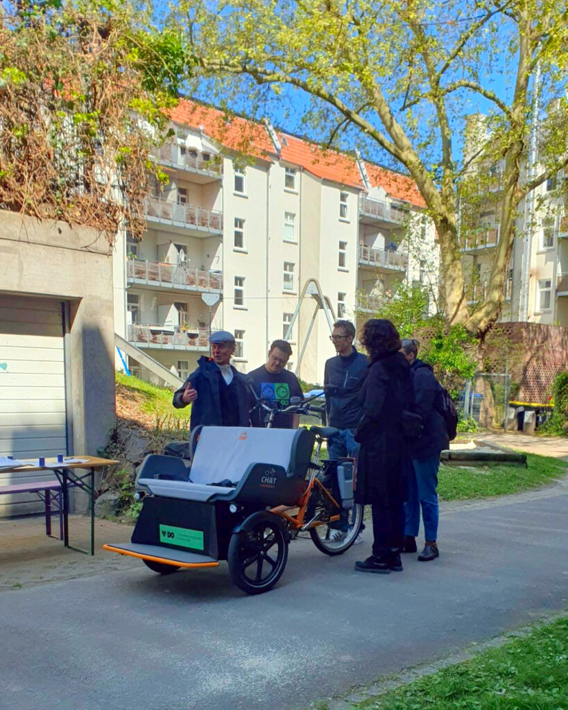 Fünf Personen stehen in einem Innenhof um eine Rikscha herum und diskutieren.