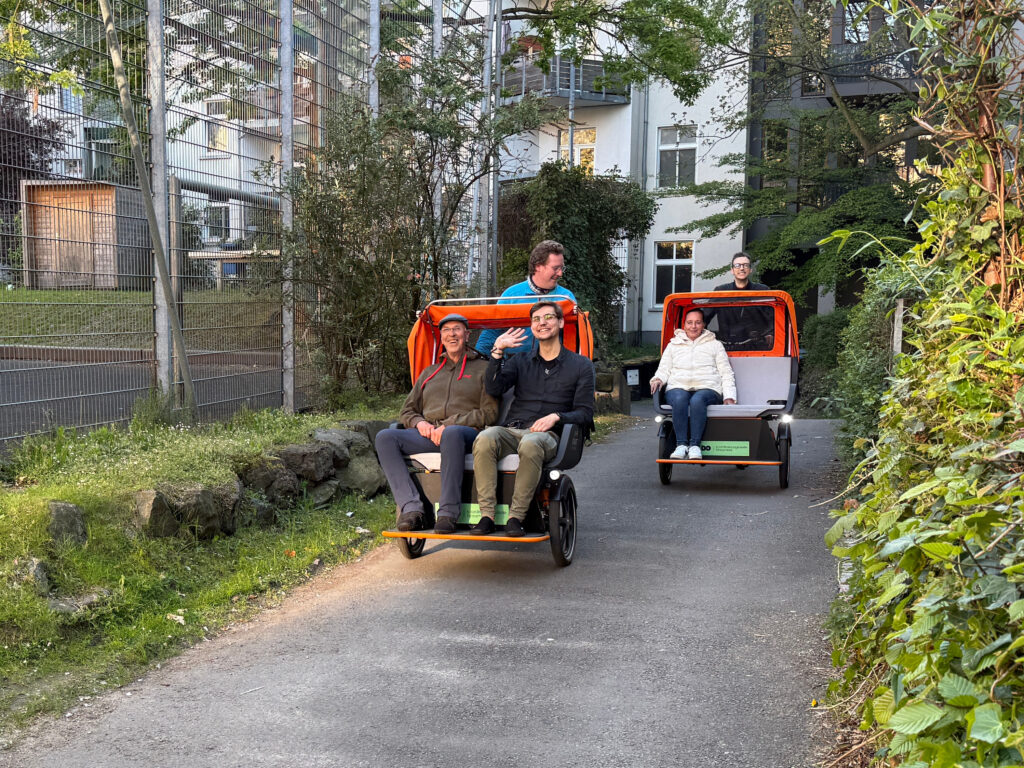 Zwei Rikschas fahren hintereinander her. Auf der vorderen Rikscha sitzen vor dem Fahrer ein junger Mann, der in die Kamera winkt, und ein lächelnder älterer Herr. Die hintere Rikscha ist neben dem Fahrer mit einer Dame besetzt.